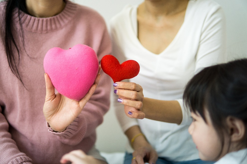 Two women holding red and pink hearts in their hands and showing it to a baby